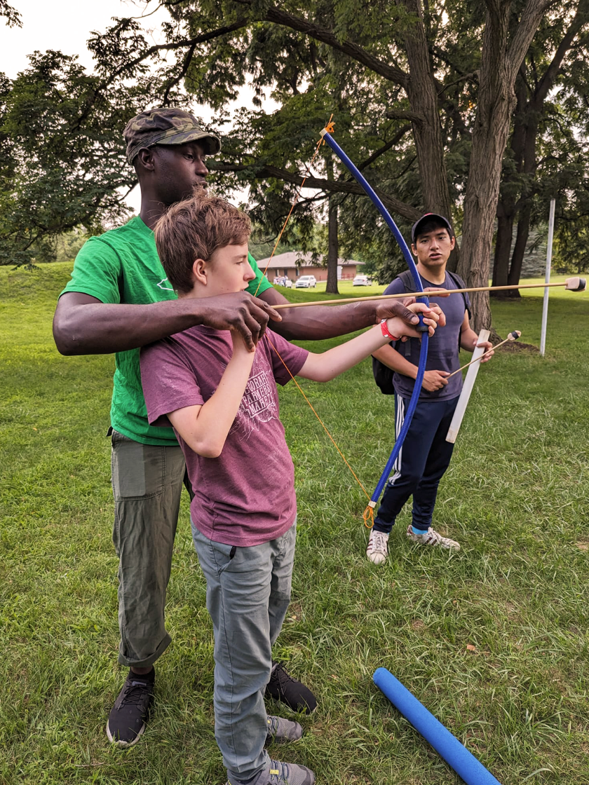 A camp counsellor helps a young boy shoot a bow and arrow
