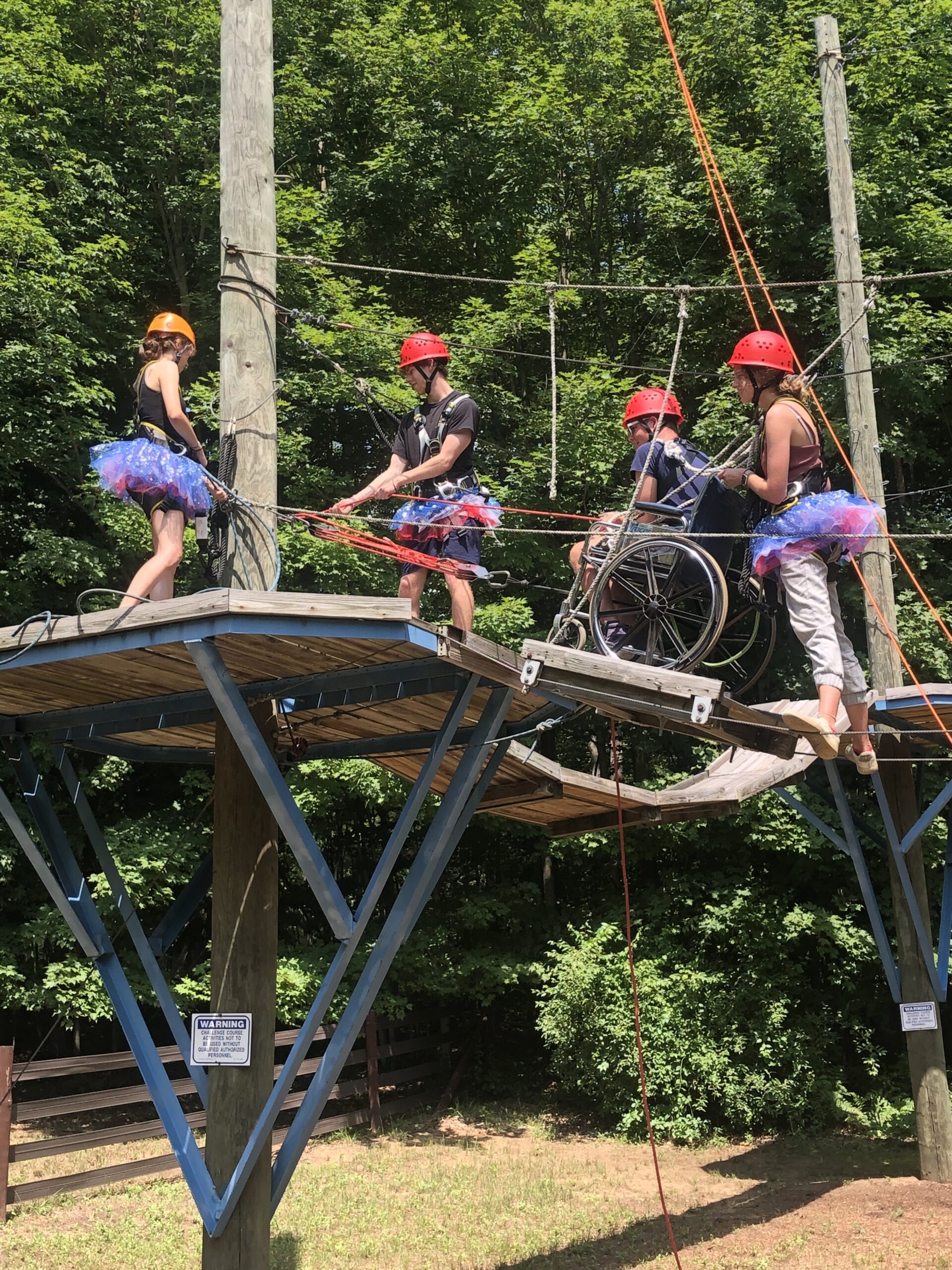 Three camp counsellors help a boy in a wheelchair across the high ropes course at The Fowler Center